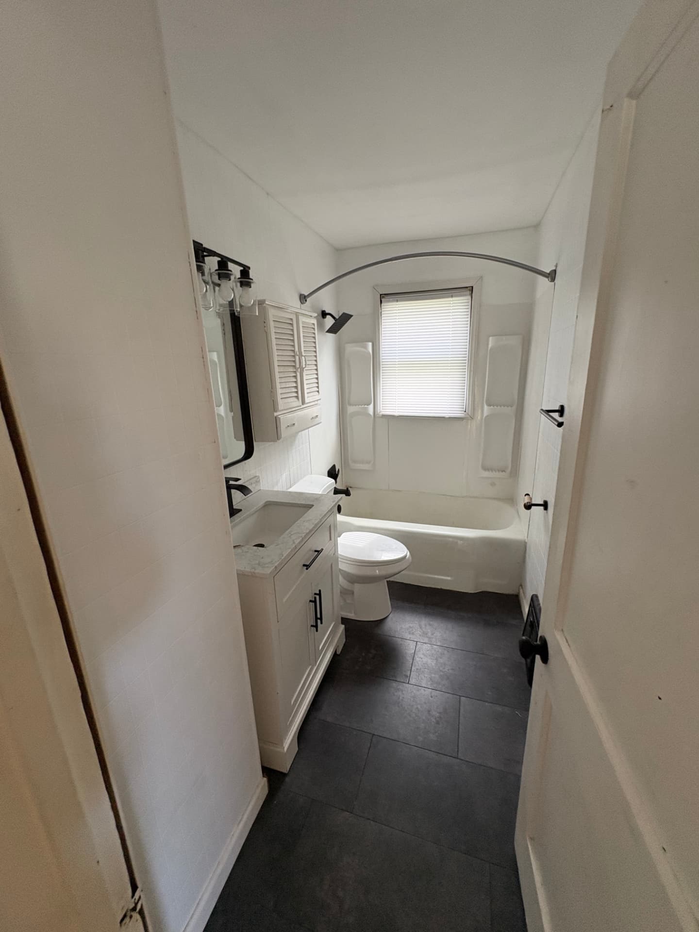 Narrow bathroom with white walls, dated fixtures and dark tile flooring before paint and deep cleaning